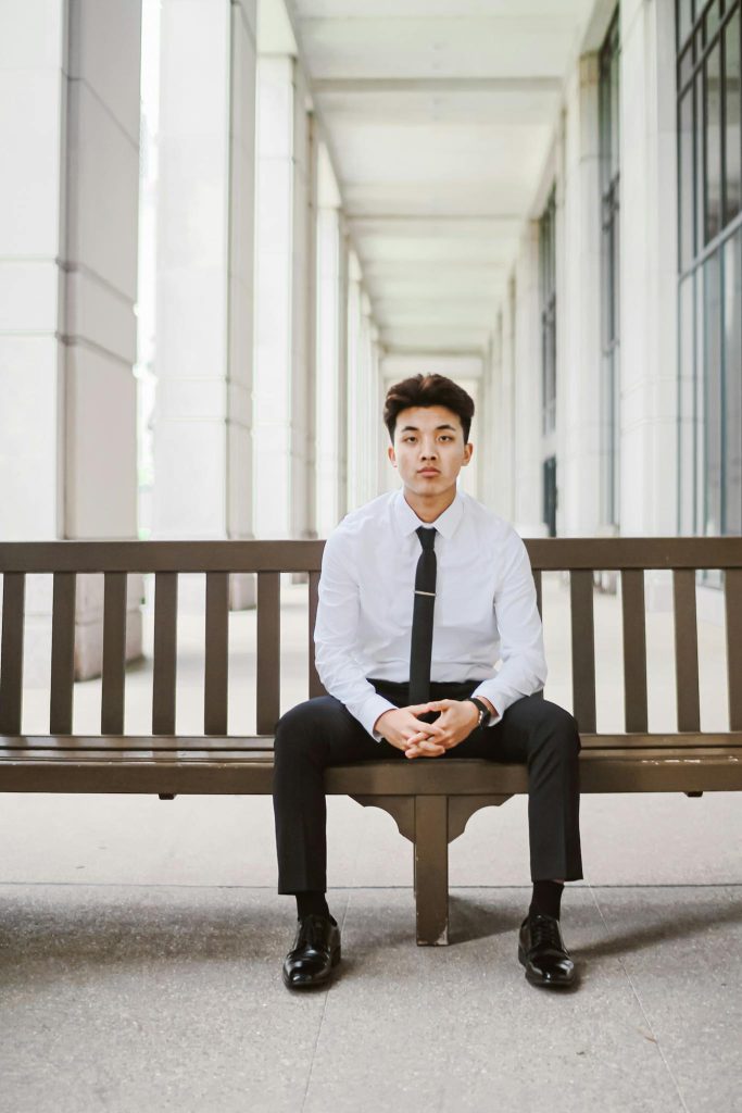 Confident young Asian man in formal attire sitting on a bench in a modern corridor.