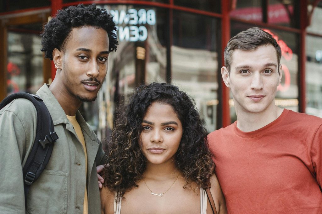 Content multiethnic group of friends looking at camera and hugging while standing on street near red modern house in city