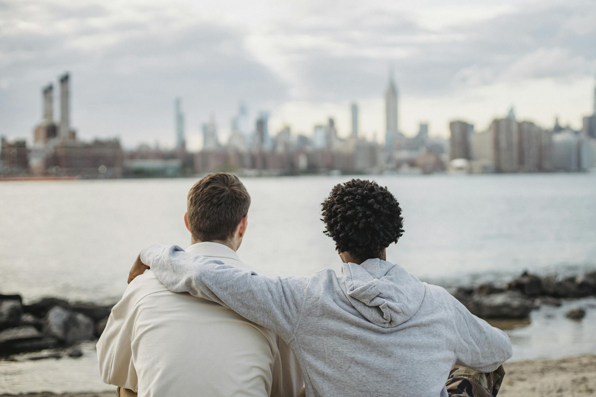 Friends sitting by the riverside, embracing with a cityscape backdrop.